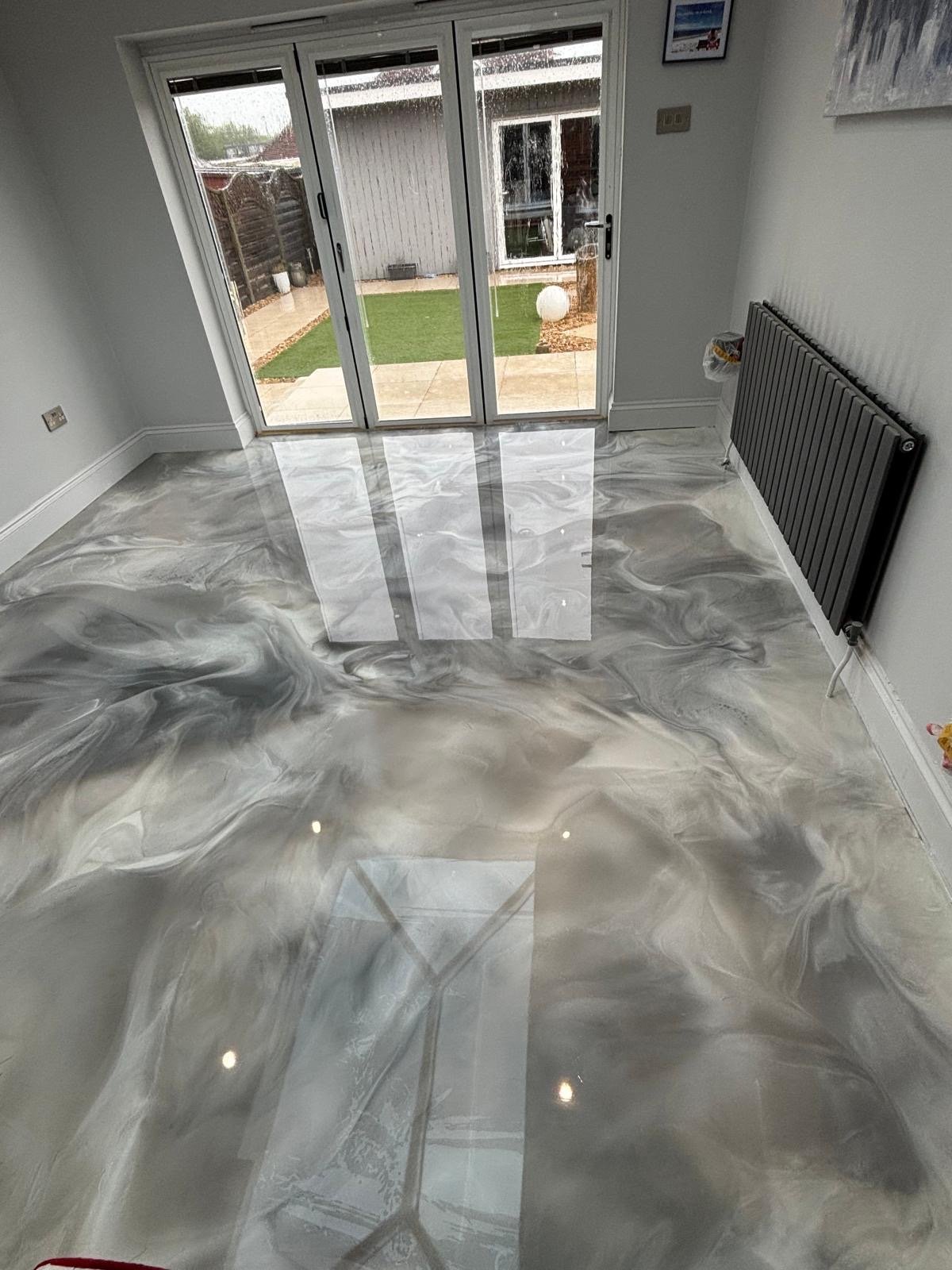 Polished epoxy floor in modern room with grey and white marble pattern, glass doors opening to garden, black radiator on right